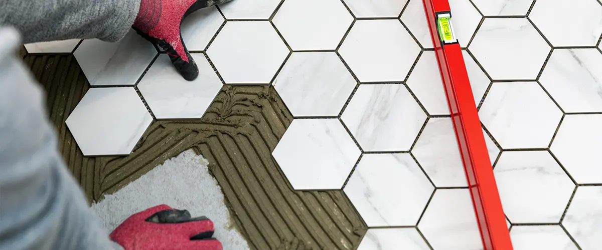 Worker carefully laying white hexagonal tiles with a red level on a tiled bathroom floor.